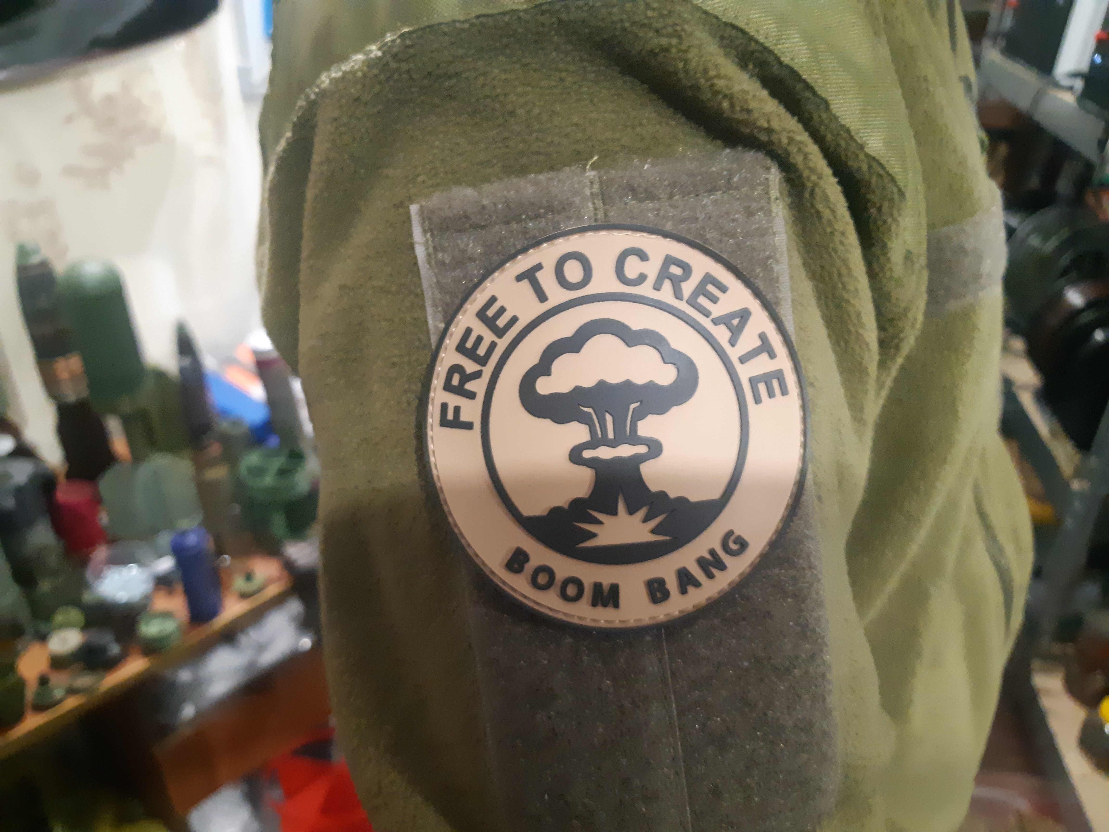 A morale patch reading „Free to Create - Boom Bang" with a mushroom-cloud emblem, and artillery munitions on shelves in the background.