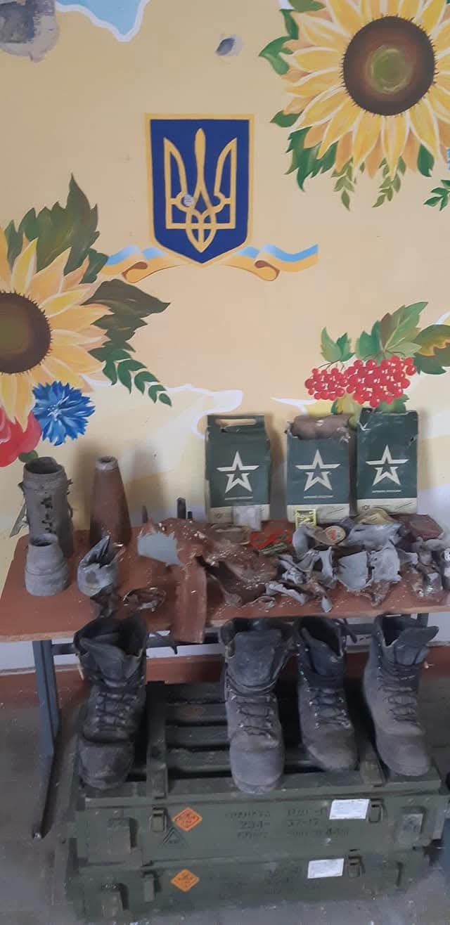 Fragments of Russian munitions and 'Russian Army' ration boxes displayed beneath a wall painted with the Ukrainian trident and sunflowers